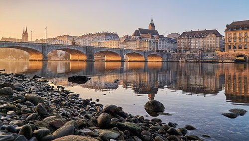 Steinbrücke über den Rhein mit historischen Gebäuden und Kirchturm in Basel bei Sonnenuntergang, Ufer mit Steinen im Vordergrund