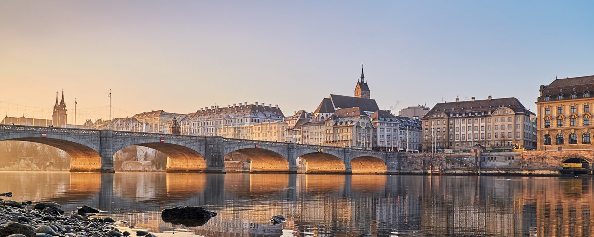 Steinbrücke über den Rhein mit historischen Gebäuden und Kirchturm in Basel bei Sonnenuntergang, Ufer mit Steinen im Vordergrund
