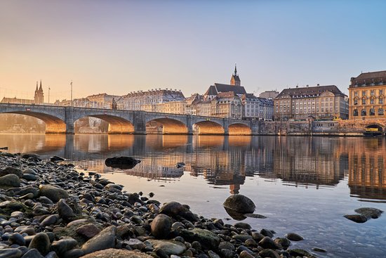Steinbrücke über den Rhein mit historischen Gebäuden und Kirchturm in Basel bei Sonnenuntergang, Ufer mit Steinen im Vordergrund