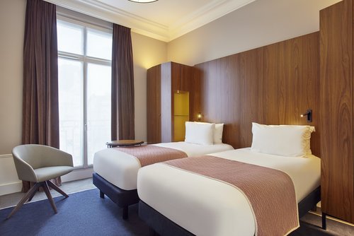 Hotelzimmer mit zwei Einzelbetten, braunen Holzpaneelen an der Wand, einem grauen Stuhl und großen Fenstern mit braunen Vorhängen.