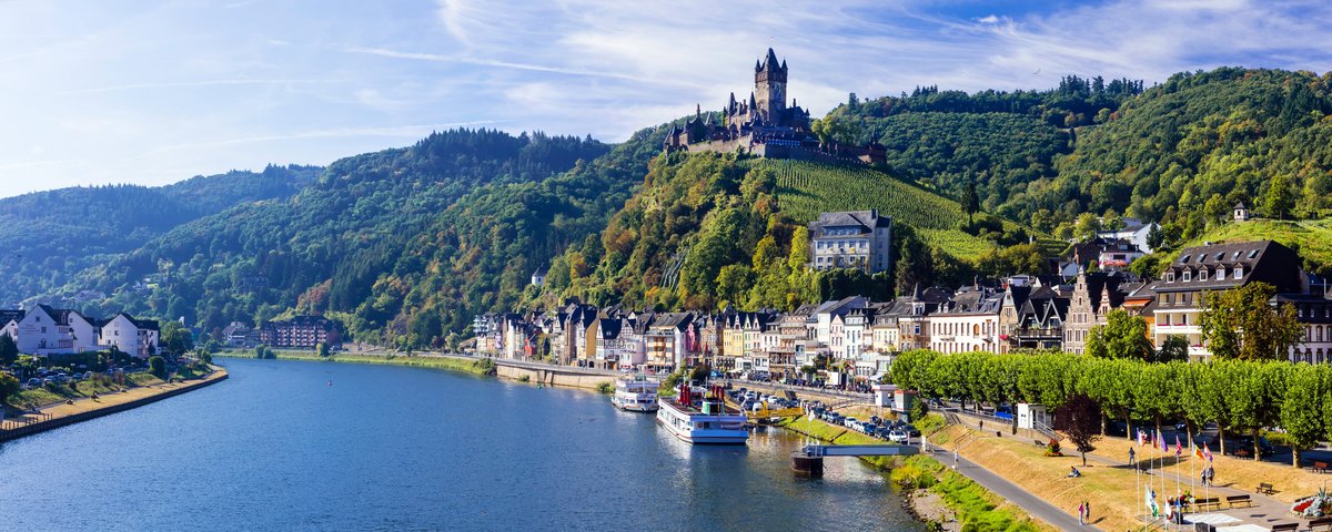Blick auf die malerische Stadt Cochem mit der Reichsburg auf einem Hügel, umgeben von grünen Weinbergen und dem Fluss Mosel im Vordergrund.