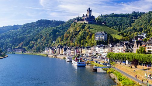 Blick auf die malerische Stadt Cochem mit der Reichsburg auf einem Hügel, umgeben von grünen Weinbergen und dem Fluss Mosel im Vordergrund.