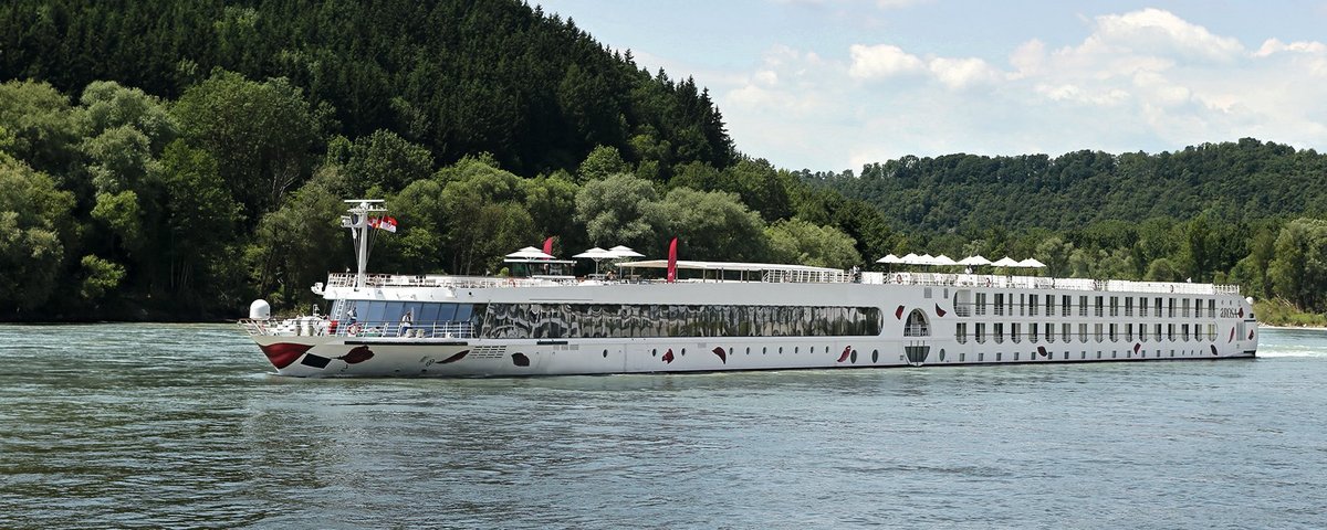 Die A-ROSA SILVA fährt am Nachmittag auf dem Rhein entlang am Ufer grüne, bewaldete Hügeln, der Himmel ist blau und leicht bewölkt.