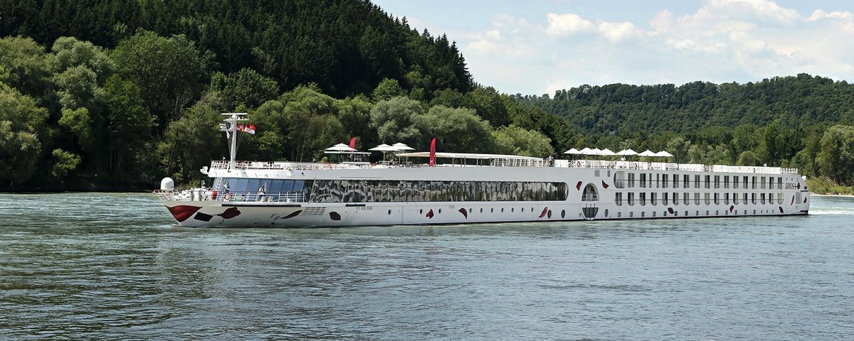 Die A-ROSA SILVA fährt am Nachmittag auf dem Rhein entlang am Ufer grüne, bewaldete Hügeln, der Himmel ist blau und leicht bewölkt.