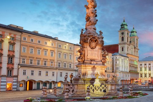 Die barocke Dreifaltigkeitssäule in Linz auf einem Platz umgeben von historischen Häusern bei Abendstimmung mit bewölktem Himmel.