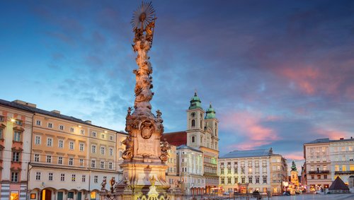 Die barocke Dreifaltigkeitssäule in Linz auf einem Platz umgeben von historischen Häusern bei Abendstimmung mit bewölktem Himmel.