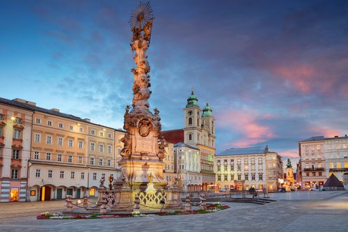 Die barocke Dreifaltigkeitssäule in Linz auf einem Platz umgeben von historischen Häusern bei Abendstimmung mit bewölktem Himmel.