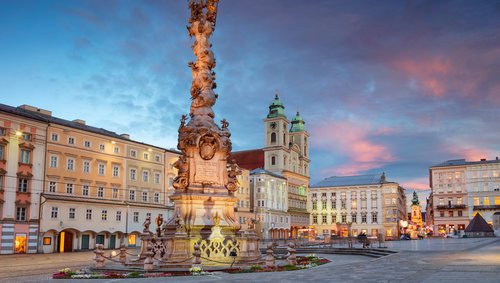 Die barocke Dreifaltigkeitssäule in Linz auf einem Platz umgeben von historischen Häusern bei Abendstimmung mit bewölktem Himmel.