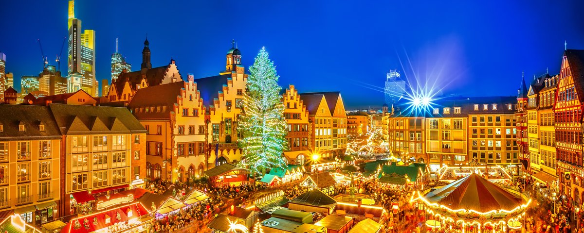 Weihnachtsmarkt in der Altstadt von Frankfurt am Main bei Nacht, mit festlich beleuchteten Ständen und einem großen, geschmückten Weihnachtsbaum und Hochhäusern im Hintergrund.