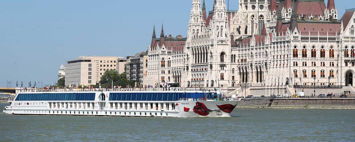 Die A-ROSA RIVA fährt auf der Donau an Budapest entlang. Viele Menschen stehen auf dem Sonnendeck, um das Parlamentsgebäude am Ufer anzusehen.