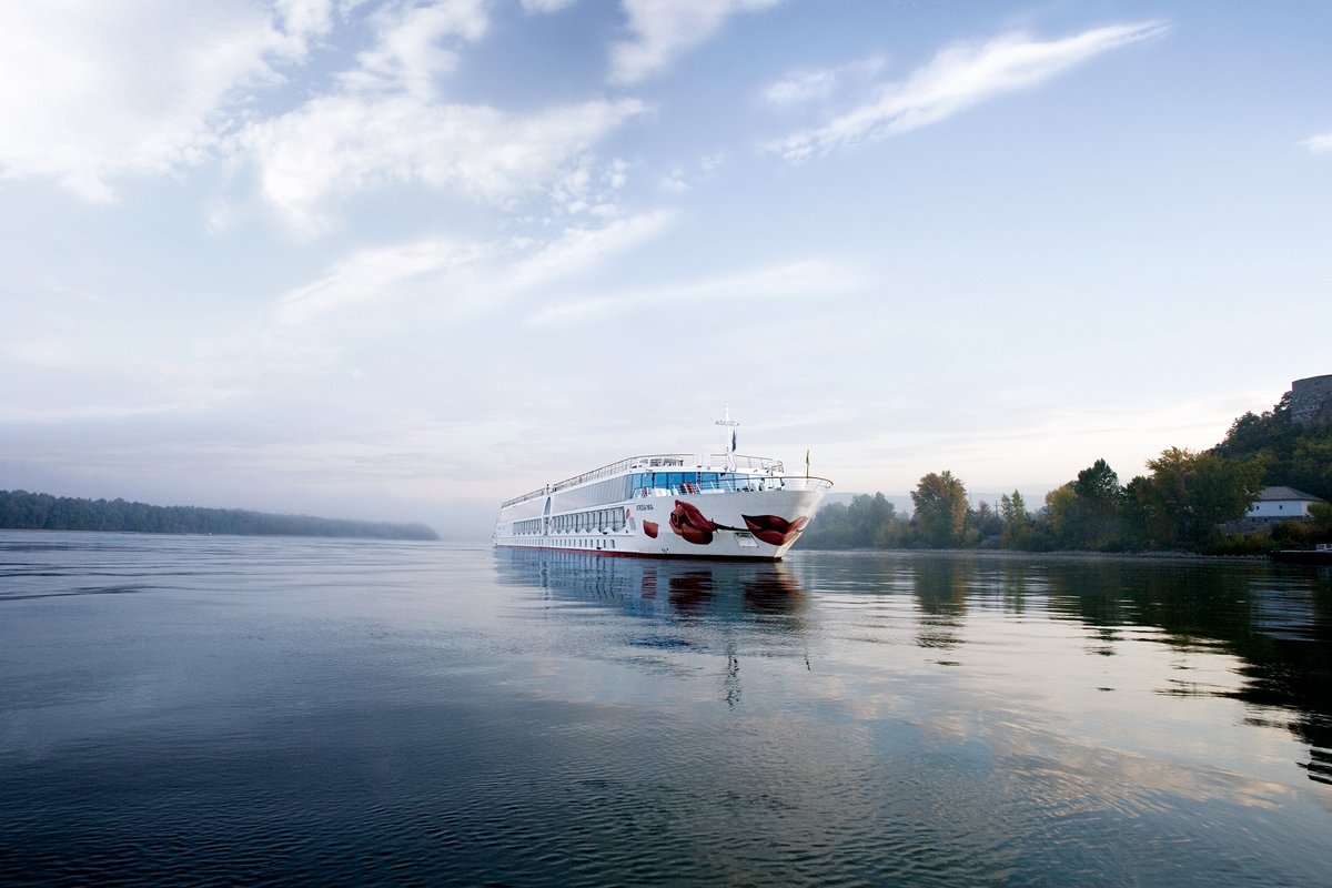 Das Flusskreuzfahrtschiff A-ROSA MIA auf der Donau. Der ruhige Fluss mit Bäumen am Ufer umgibt das Schiff mit dem markanten Kussmund un der Rose auf dem Bug.