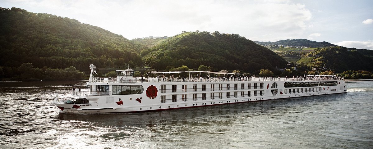 Ein Schiff der A-ROSA Rheinflotte gleitet am Nachmittag auf dem Rhein entlang, am Ufer grüne, bewaldete Hügeln und kleinen Siedlungen im Hintergrund.