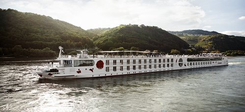 Ein Schiff der A-ROSA Rheinflotte gleitet am Nachmittag auf dem Rhein entlang, am Ufer grüne, bewaldete Hügeln und kleinen Siedlungen im Hintergrund.