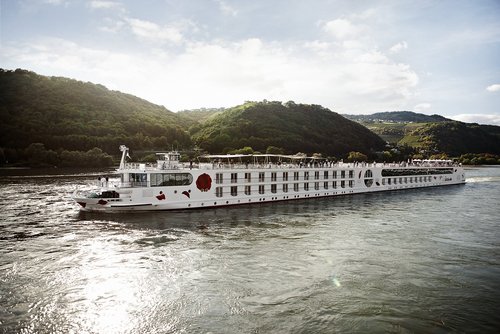 Ein Schiff der A-ROSA Rheinflotte gleitet am Nachmittag auf dem Rhein entlang, am Ufer grüne, bewaldete Hügeln und kleinen Siedlungen im Hintergrund.