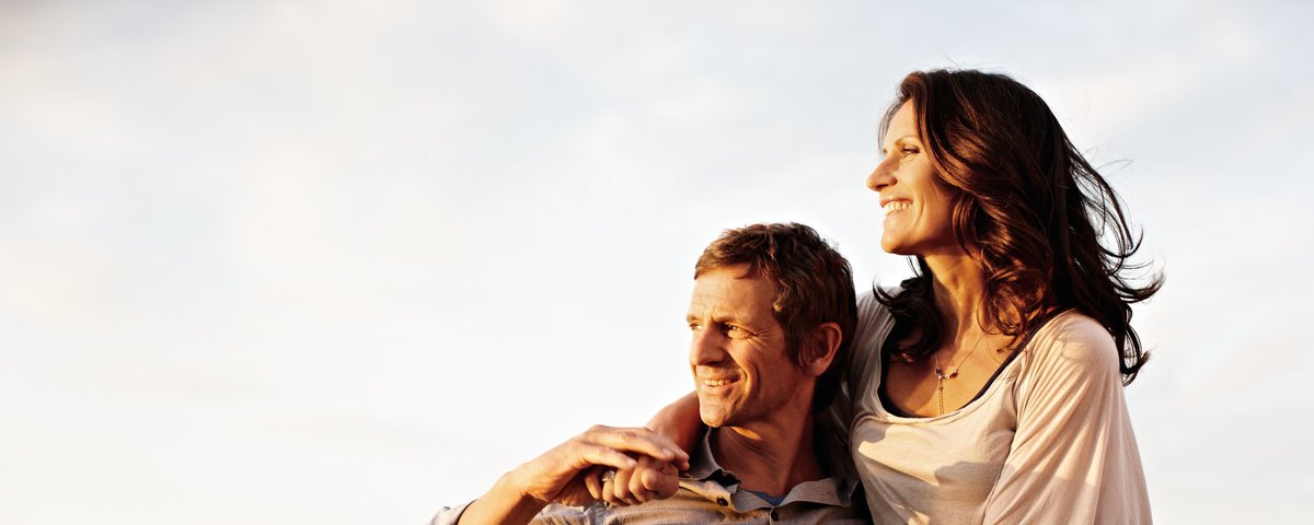 Eine Frau sitzt auf der Reling eines Flusskreuzfahrtschiffs und legt die Arme auf den Mann neben ihr, der sich an die Reling lehnt und sie festhält. Sie genießen den Blick auf den Rhein bei Sonnenschein.