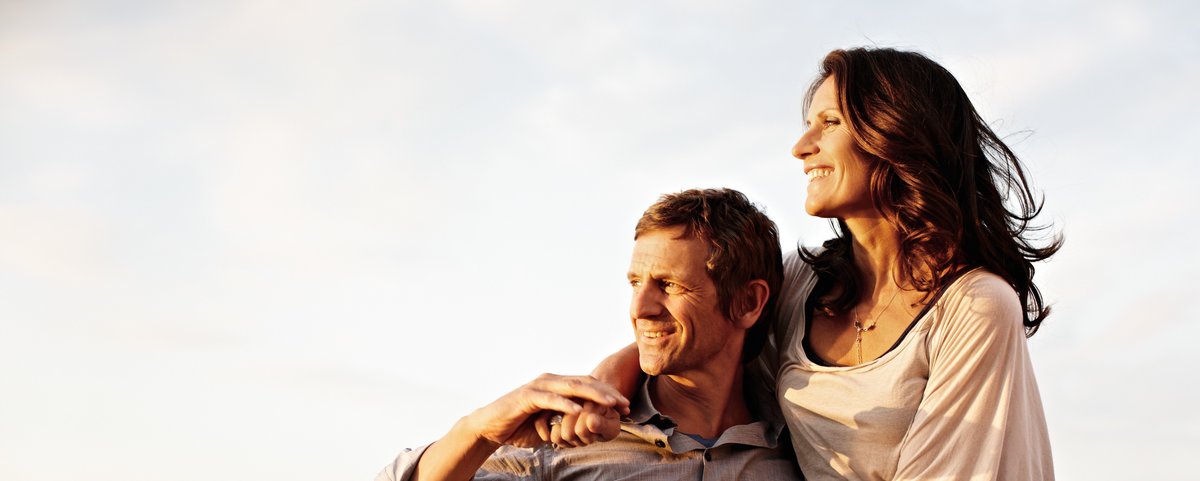 Eine Frau sitzt auf der Reling eines Flusskreuzfahrtschiffs und legt die Arme auf den Mann neben ihr, der sich an die Reling lehnt und sie festhält. Sie genießen den Blick auf den Rhein bei Sonnenschein.