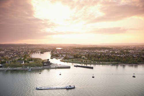 Panoramablick auf die Stadt Koblenz mit dem Deutschen Eck. Auf dem Wasser fahren Boote und im Vordergrund ist eine Hochseilbahn unter einem strahlenden Himmel bei Sonnenuntergang. 