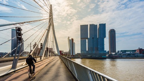 Fahrradfahrer auf der Erasmusbrücke in Rotterdam mit Blick auf moderne Hochhäuser am Flussufer bei bewölktem Himmel
