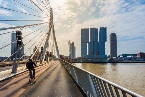 Fahrradfahrer auf der Erasmusbrücke in Rotterdam mit Blick auf moderne Hochhäuser am Flussufer bei bewölktem Himmel
