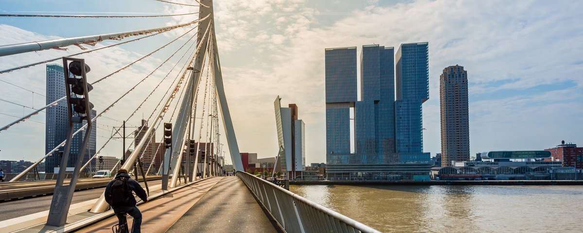 Fahrradfahrer auf der Erasmusbrücke in Rotterdam mit Blick auf moderne Hochhäuser am Flussufer bei bewölktem Himmel