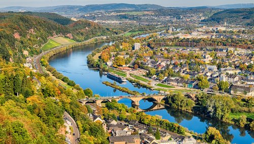 Blick auf die Stadt Trier mit der Mosel, einer historischen Steinbrücke und herbstlich gefärbten Bäumen entlang des Flusses