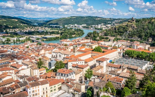 Blick auf die Stadt Vienne in Frankreich mit der Rhône im Hintergrund, umgeben von grünen Hügeln und historischen Gebäuden und einer Burgruine auf einem Hügel