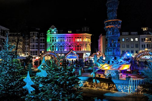 Weihnachtsmarkt in Koblenz bei Nacht mit bunt beleuchtetem historischen Gebäude, geschmückten Tannenbäumen und einem beleuchteten Brunnen mit Figuren