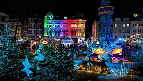 Weihnachtsmarkt in Koblenz bei Nacht mit bunt beleuchtetem historischen Gebäude, geschmückten Tannenbäumen und einem beleuchteten Brunnen mit Figuren