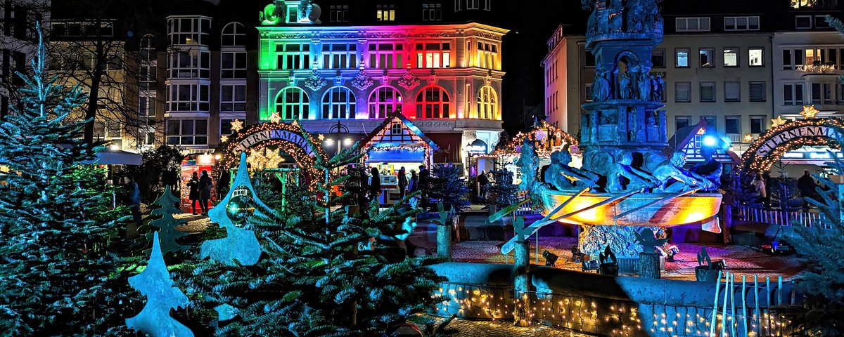 Weihnachtsmarkt in Koblenz bei Nacht mit bunt beleuchtetem historischen Gebäude, geschmückten Tannenbäumen und einem beleuchteten Brunnen mit Figuren