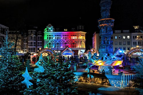 Weihnachtsmarkt in Koblenz bei Nacht mit bunt beleuchtetem historischen Gebäude, geschmückten Tannenbäumen und einem beleuchteten Brunnen mit Figuren