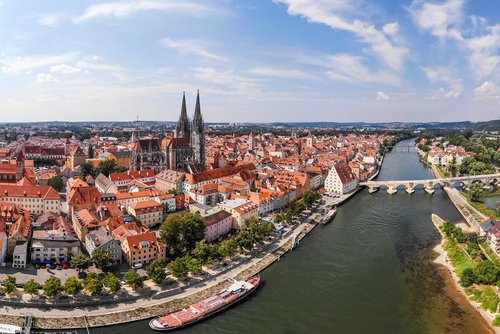 Sommerliches Panorama der Regensburger Altstadt. Im Vordergrund bahnt sich die Donau durch die Stadt. Im Hintergrund sind historische Wohngebäude und eine Kathedrale.