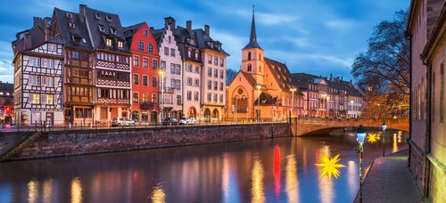 Abendliche Ansicht der historischen Fachwerkhäuser und einer Kirche am Flussufer in Straßburg mit beleuchteten Straßenlaternen und Reflexionen im Wasser