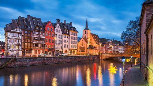 Abendliche Ansicht der historischen Fachwerkhäuser und einer Kirche am Flussufer in Straßburg mit beleuchteten Straßenlaternen und Reflexionen im Wasser