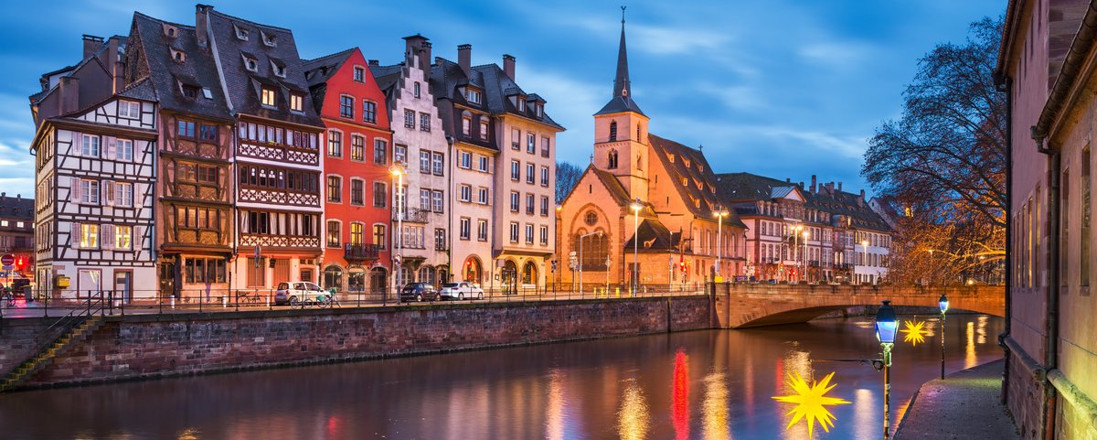 Abendliche Ansicht der historischen Fachwerkhäuser und einer Kirche am Flussufer in Straßburg mit beleuchteten Straßenlaternen und Reflexionen im Wasser