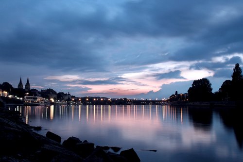 Blick auf die Stadt Koblenz in der Abenddämmerung unter einem bewölkten Himmel. Im Vordergrund der Fluss, in dem sich die Lichter der Stadt spiegeln.