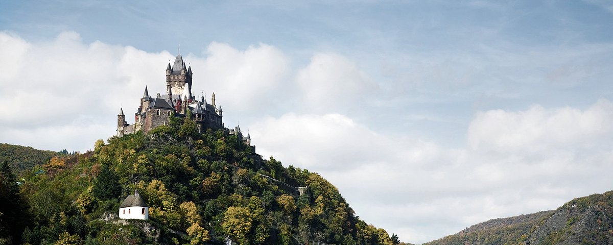 Blick auf die Reichsburg Cochem, umgeben von grünen Hügeln und Cochem an der Mosel mit Schwänen, unter einem bewölkten blauen Himmel.