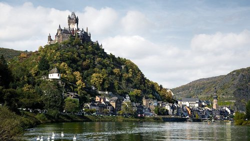 Blick auf die Reichsburg Cochem, umgeben von grünen Hügeln und Cochem an der Mosel mit Schwänen, unter einem bewölkten blauen Himmel.