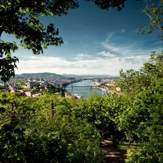 Panoramablick auf Budapest von einem grün bewachsenen Park aus auf die Brücke über der Donau und die Stadt mit Hügellandschaft im Hintergrund.