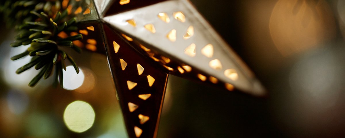 Metallstern mit kleinen dreieckigen Aussparungen, der warmes Licht ausstrahlt, neben Tannenzweigen in unscharfem Hintergrund.