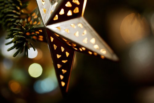 Metallstern mit kleinen dreieckigen Aussparungen, der warmes Licht ausstrahlt, neben Tannenzweigen in unscharfem Hintergrund.