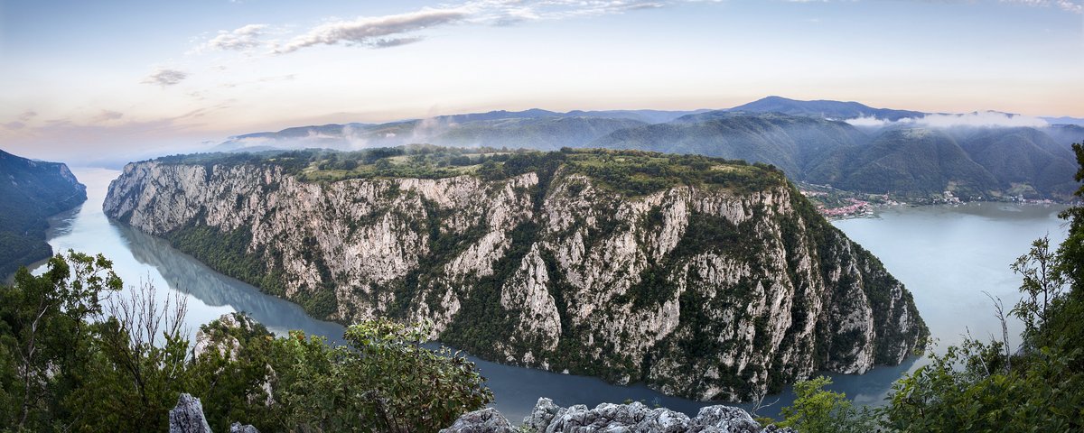 Panoramablick auf die Kataraktenstrecke der Donau mit steilen bewachsenen Felsen und einem leicht bewölktem Himmel, im Hintergrund eine Ortschaft am Fluss und Hügellandschaft.