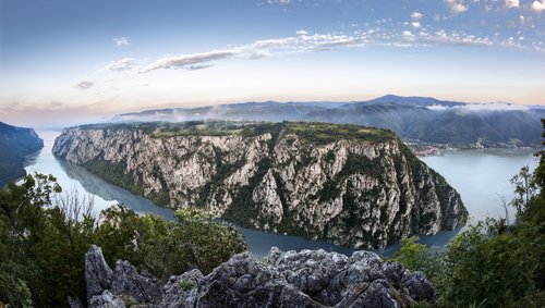 Panoramablick auf die Kataraktenstrecke der Donau mit steilen bewachsenen Felsen und einem leicht bewölktem Himmel, im Hintergrund eine Ortschaft am Fluss und Hügellandschaft.