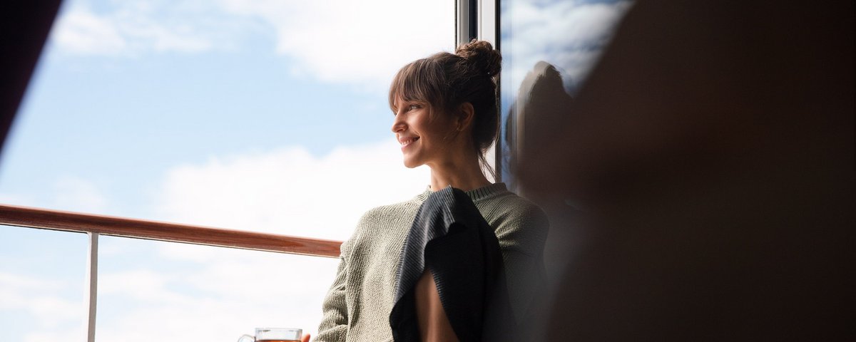 Eine Frau sitzt entspannt mit einem Glas Tee in der Hand in einer Kabine auf einem Flusskreuzfahrtschiff vor dem französischen Balkon und schaut bei geöffneter Tür hinaus auf die Donau.