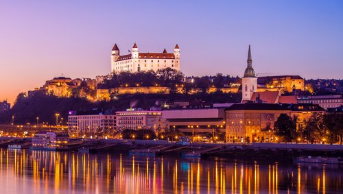 Blick auf das Donauufer bei Bratislava bei Sonnenuntergang mit der Burg Bratislava im Hintergrund, umgeben von Bäumen und der Stadt. Im Vordergrund spiegeln sich die Lichter im Fluss.