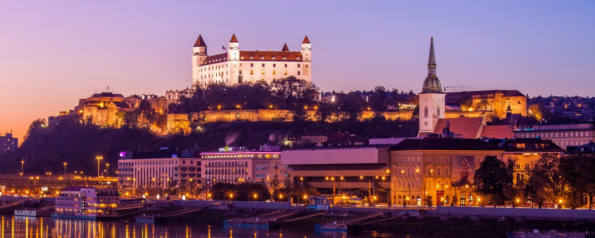 Blick auf das Donauufer bei Bratislava bei Sonnenuntergang mit der Burg Bratislava im Hintergrund, umgeben von Bäumen und der Stadt. Im Vordergrund spiegeln sich die Lichter im Fluss.