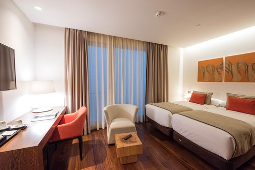 Hotelzimmer mit zwei Einzelbetten, braunen Bettläufern und orangen Kissen, Schreibtisch mit rotem Stuhl, Fernseher an der Wand und einem kleinen weißen Sessel vor einem Fenster mit Vorhängen.