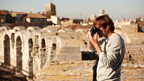 Ein Mann steht auf einer historischen Mauer in Arles und fotografiert etwas. Im Hintergrund die gelben Gebäude der Stadt.