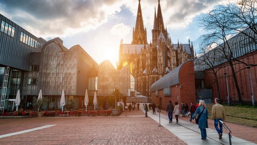 Rückansicht des Kölner Doms bei Sonnenuntergang und mordernen Gebäuden im Vordergrund. Spaziergänger passieren die Gebäude