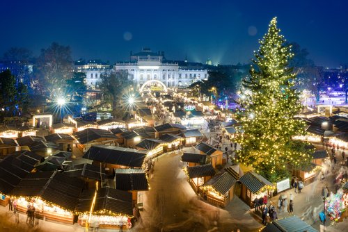Blick auf den Wiener Weihnachtsmarkt bei Nacht. Im Hintergrund sind historische Gebäude, im Vordergrund sind Weihnachtsmarkthütten und ein festlich beleuchteter Tannenbaum.