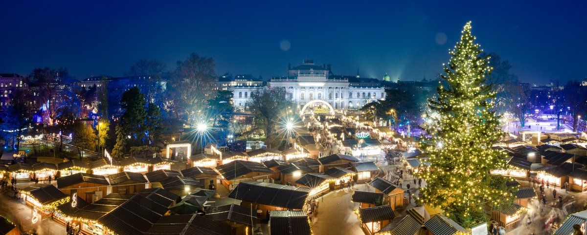 Blick auf den Wiener Weihnachtsmarkt bei Nacht. Im Hintergrund sind historische Gebäude, im Vordergrund sind Weihnachtsmarkthütten und ein festlich beleuchteter Tannenbaum.